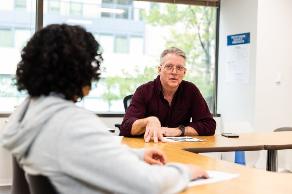 Dale Baker, 
a Sycamore School Humanities Teacher, speaks energetically with a high school student at a classroom table.