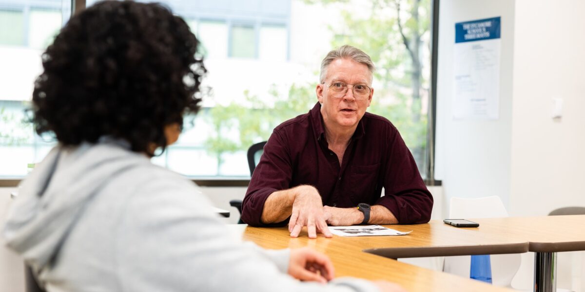 Dale Baker, a Sycamore School Humanities Teacher, speaks energetically with a high school student at a classroom table.