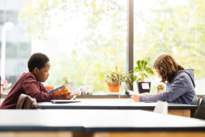 two students reading books as a table, while the light shines in through the window behind them.
