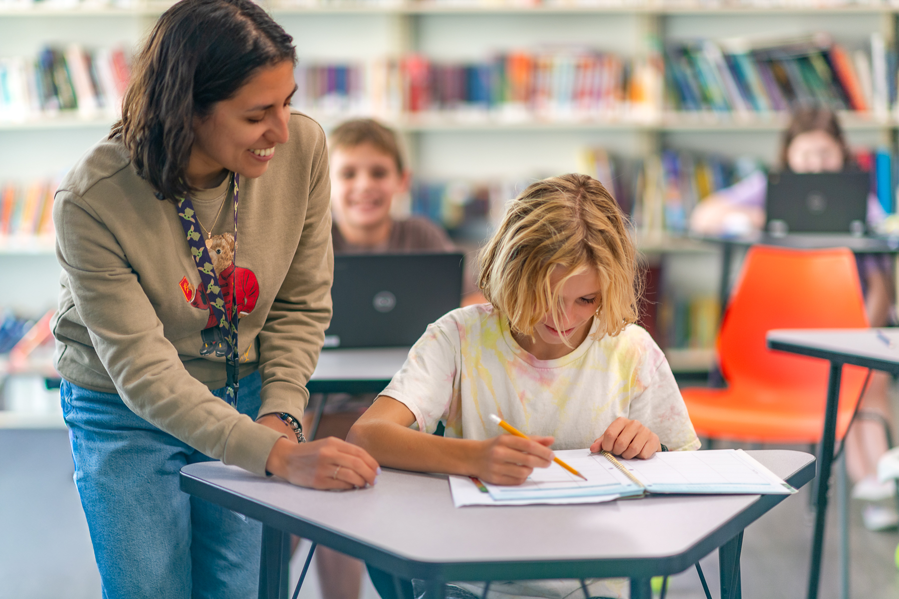 A teacher smiles while she helps her focused student work through the worksheet on their desk.
